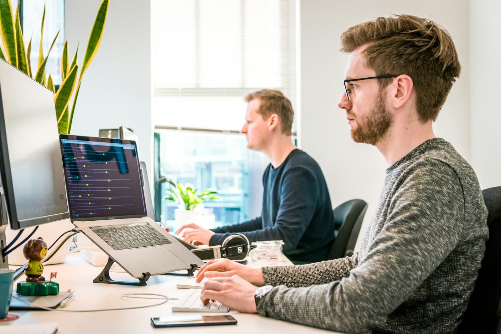 Two developers at their desks, one with a terminal open on a laptop beside a large monitor
